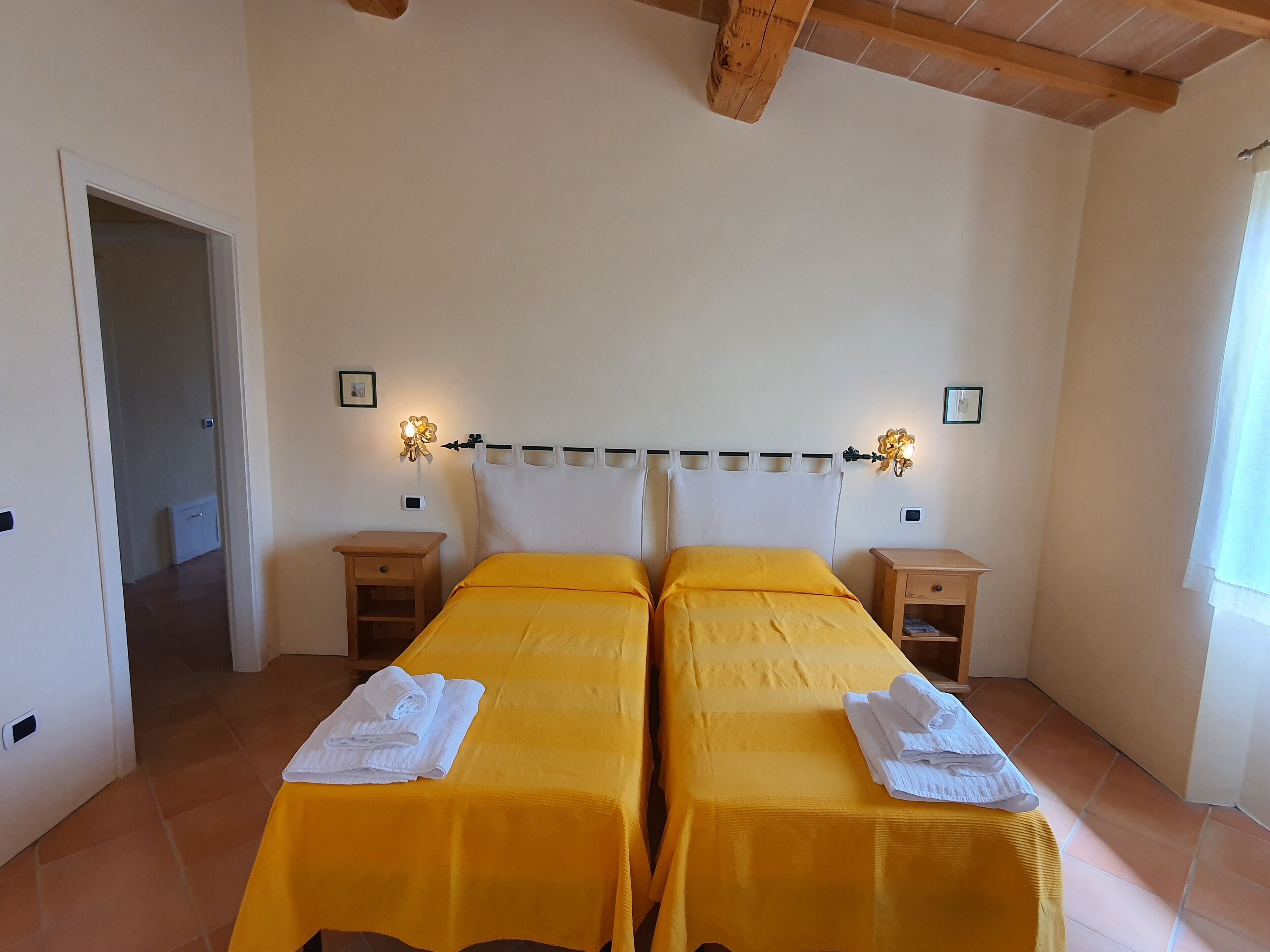Una stanza con due letti, ognuno con copriletto giallo e cuscini bianchi, affiancati da comodini. Sono presenti lampade a parete con decorazioni floreali.