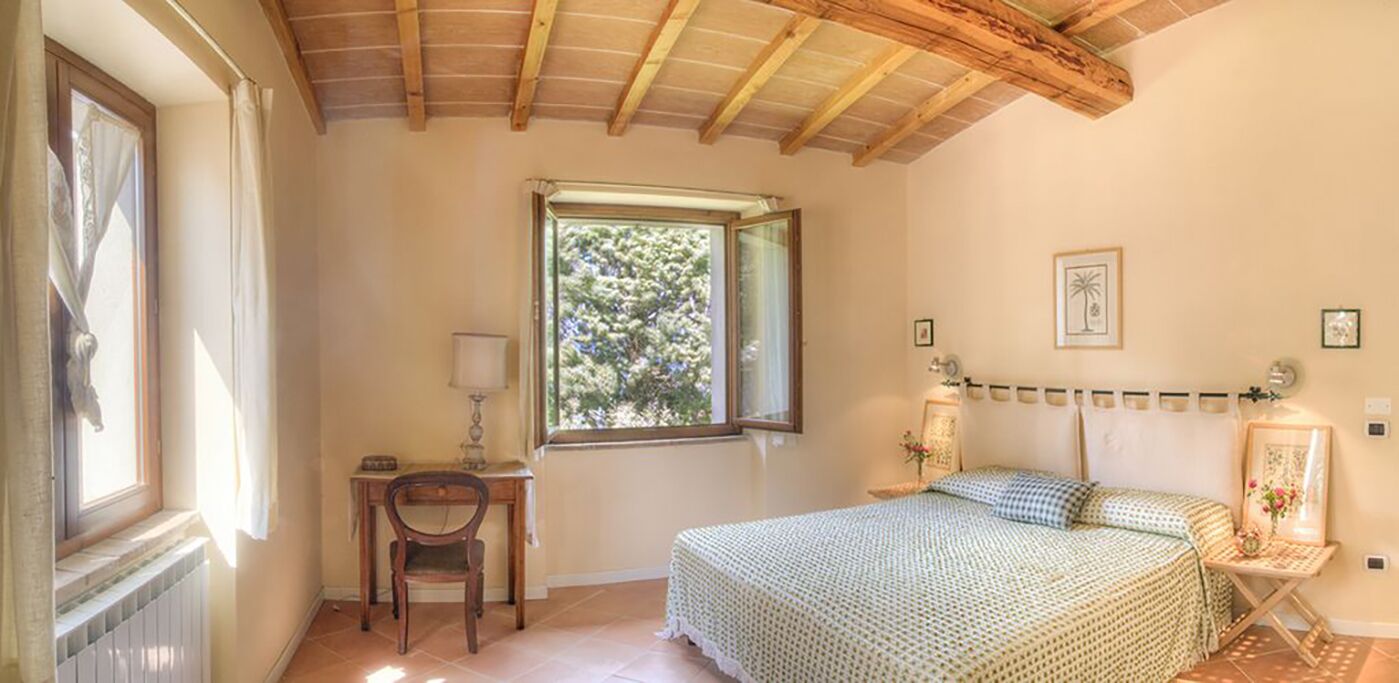 Een slaapkamer met een houten plafond, een bed met een bedspreide met patroon, een bureau met stoel, en een raam met uitzicht op bomen.