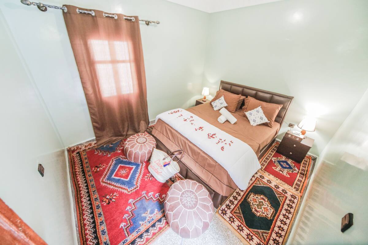 A bedroom with a bed, pillows, a nightstand, a lamp, and a window with curtains.