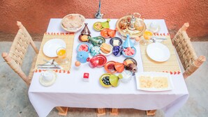 A table set with various dishes and drinks, surrounded by wicker chairs, against a textured wall. - Dar Widad - Hostel (Tarmigt)