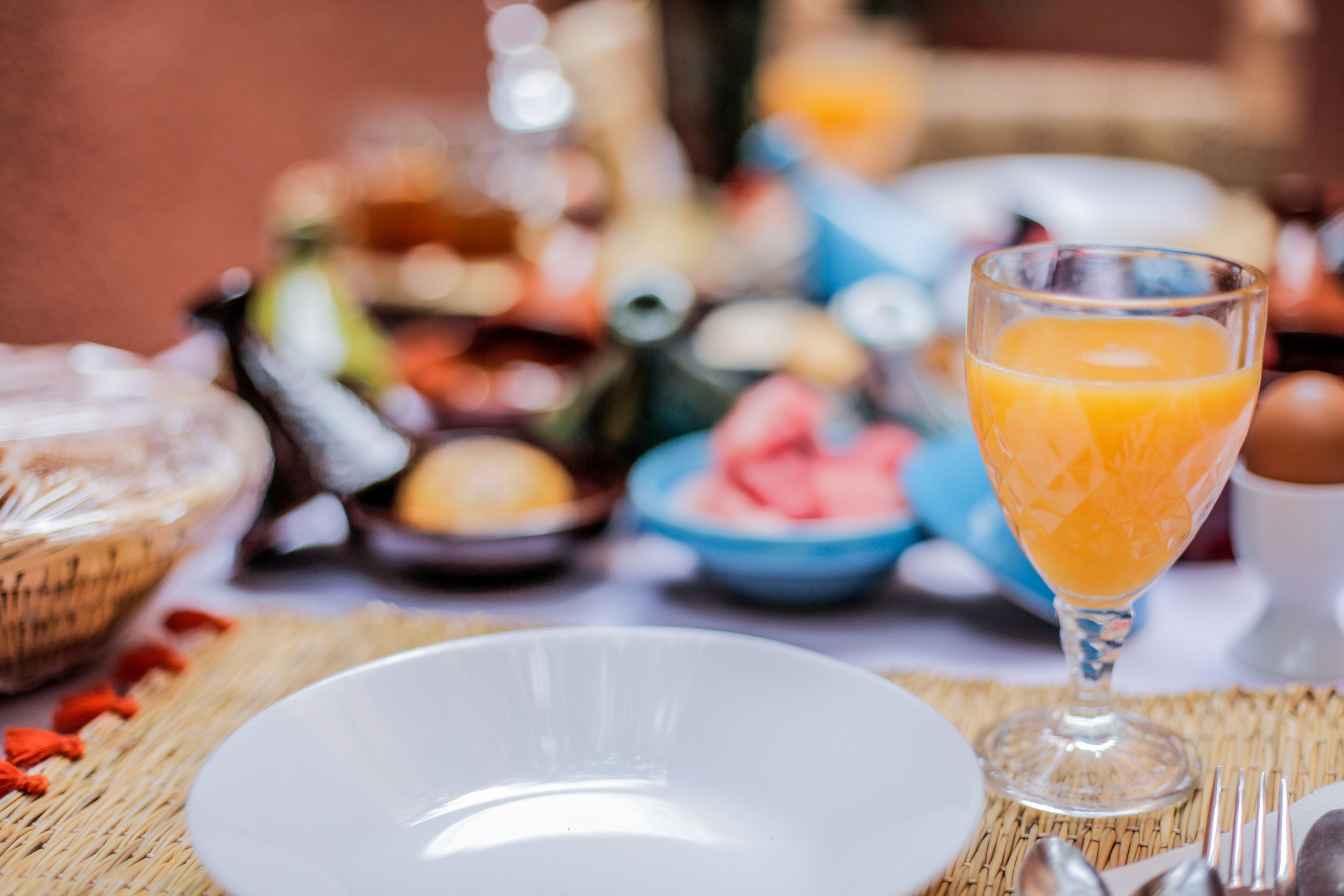 Une table mise pour le petit-déjeuner, avec un verre de jus d’orange, une assiette blanche et divers aliments en arrière-plan.