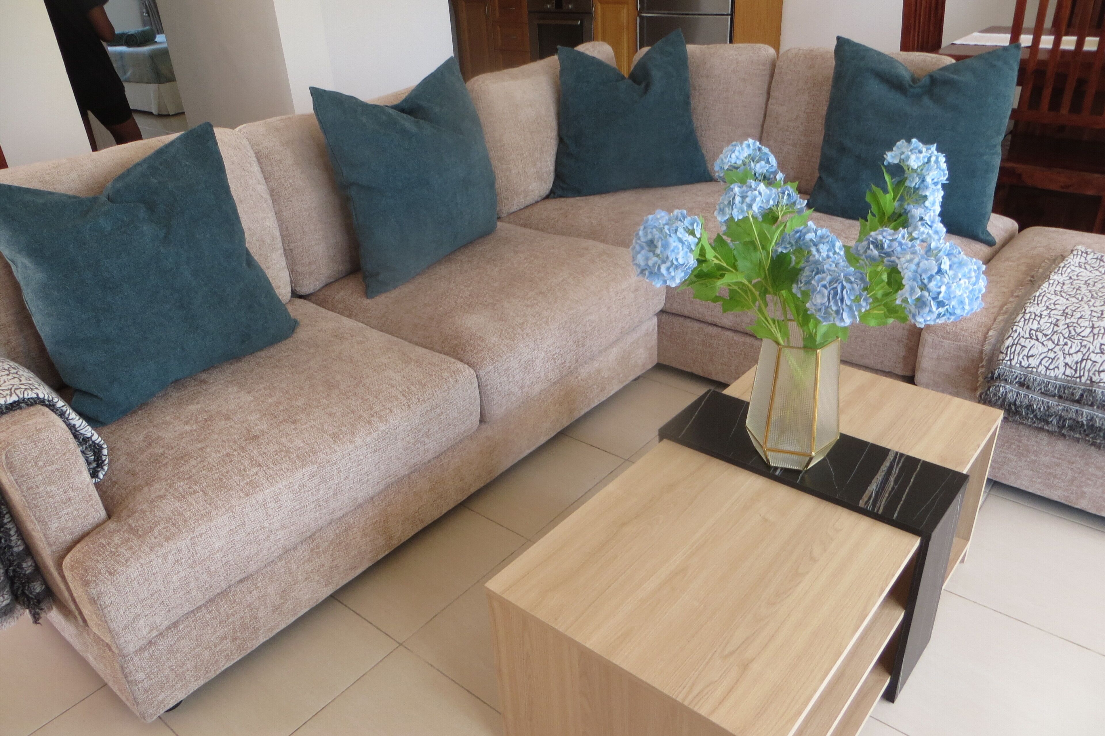 Un canapé d’angle beige avec des coussins turquoise, une table basse en bois et un vase avec des fleurs bleues.