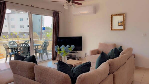 A living room with a sofa, a TV, a ceiling fan, and a balcony with chairs and a table. - Luxury G/Floor Apt, Pool, A/C, TV, WIFI, Patio/Garden & Pvt. Parking at door. (Kato Paphos)