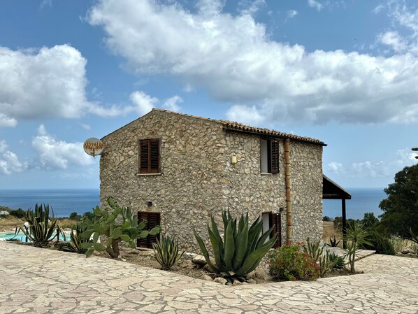 A stone house with a tiled patio, surrounded by cacti and other desert plants, overlooking a body of water.