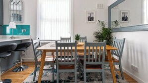 Een eetgedeelte met een houten tafel, grijze stoelen, een zwart-wit tapijt en een turquoise spatwand. Er is ook een raam met zonwering.