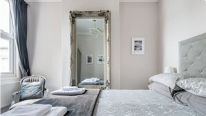 Een slaapkamer met een grote, versierde spiegel, een bed met beddengoed met een patroon, een stoel met een kussen, en een nachtkastje met opgevouwen handdoeken.