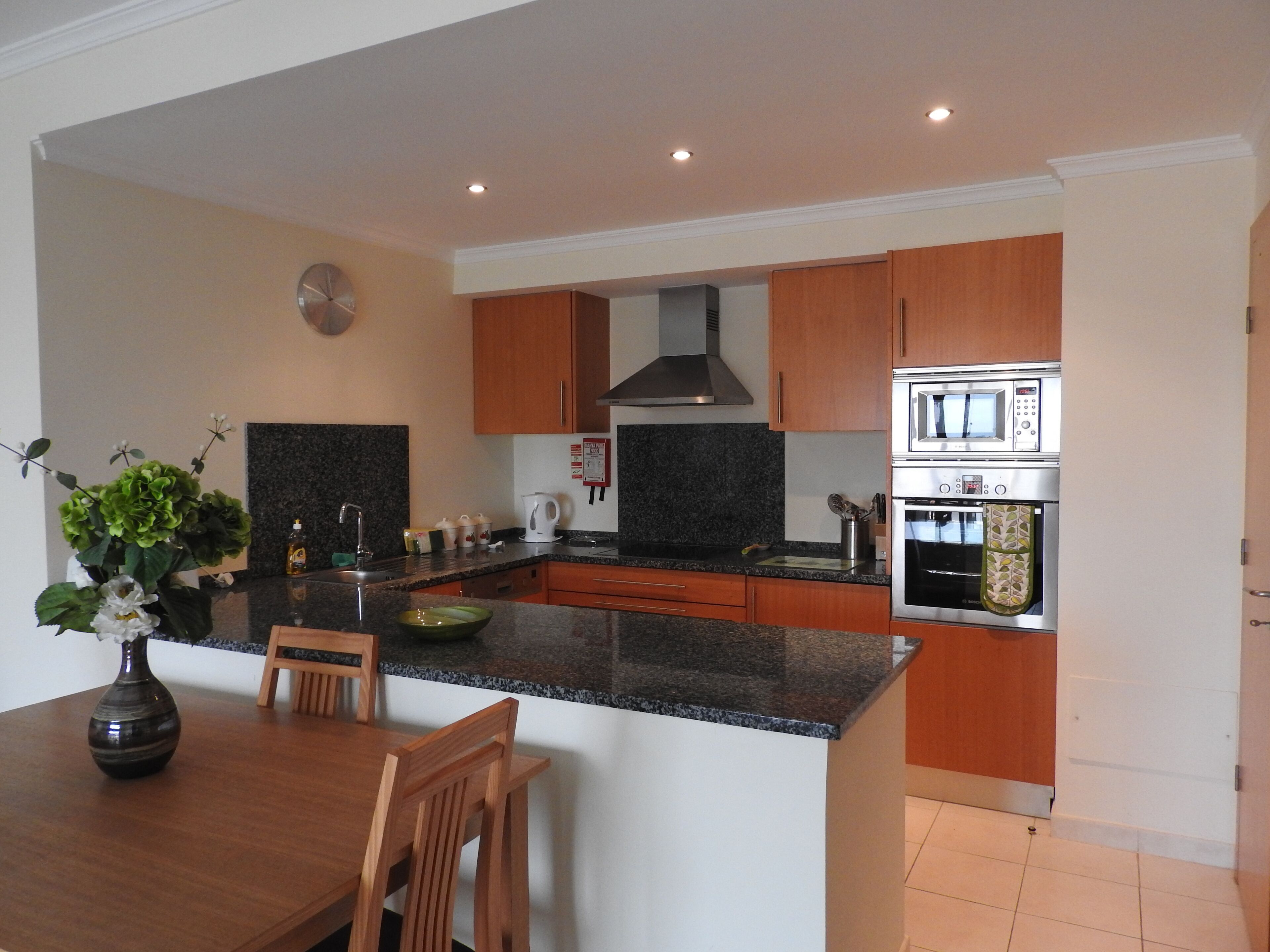 A modern kitchen with granite countertops, wooden cabinets, a built-in oven, and a dining area with a wooden table and chairs.