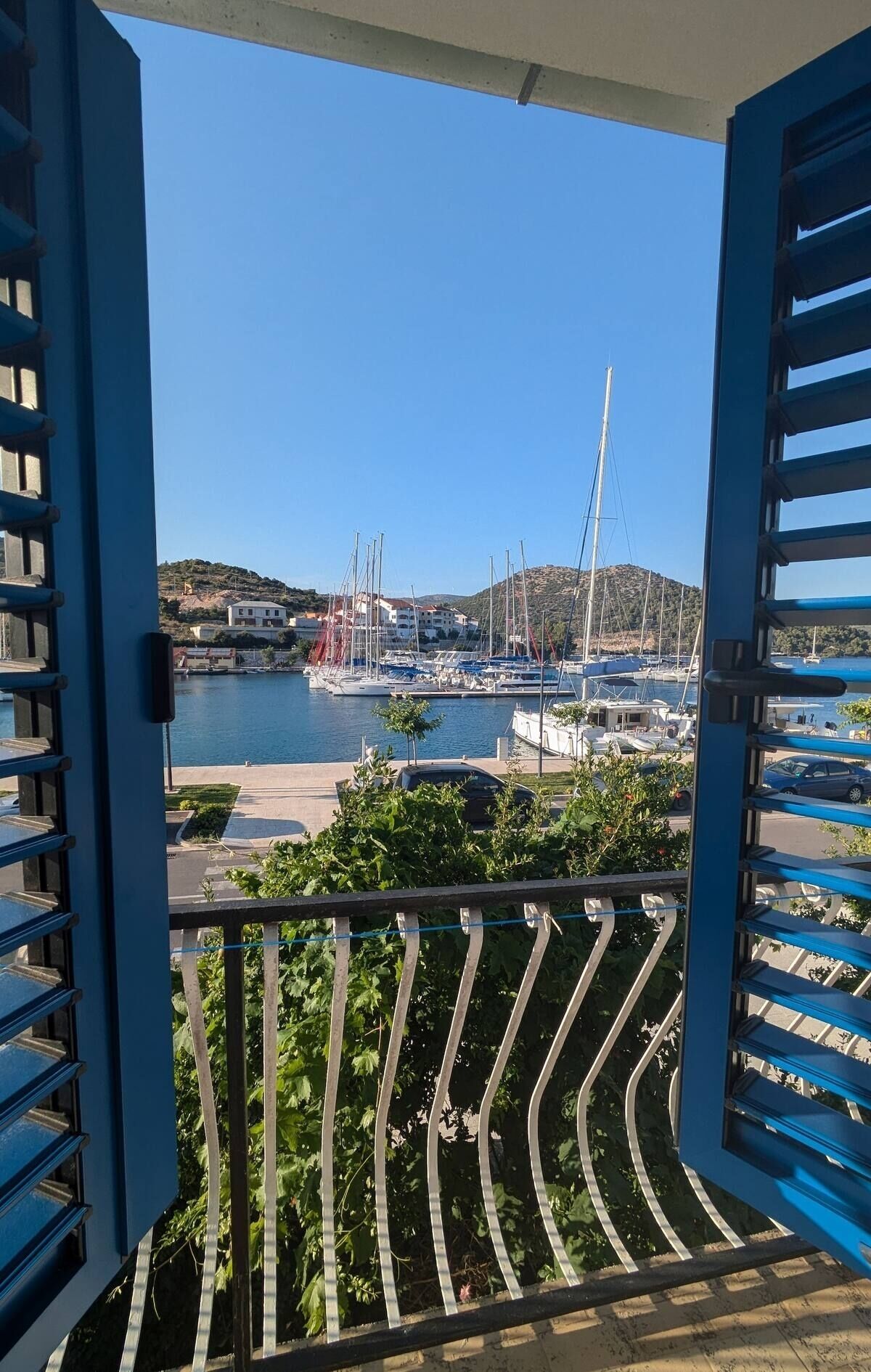 A balcony with blue shutters overlooking a marina with boats and buildings in the background.