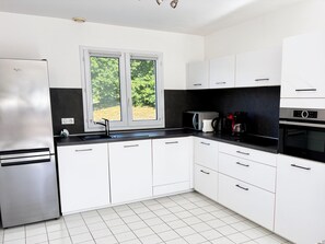 A modern kitchen with white cabinets, a stainless steel refrigerator, a double sink, and a window with a view of greenery.