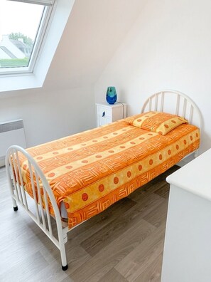 A small, single-bed room with a sloped ceiling, a white metal bed frame, a wooden floor, a small bedside table with a blue vase, and a window with a view of buildings outside.