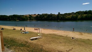 Una playa junto al lago, con gente nadando y tomando el sol, una silla de salvavidas y un bote en la orilla.