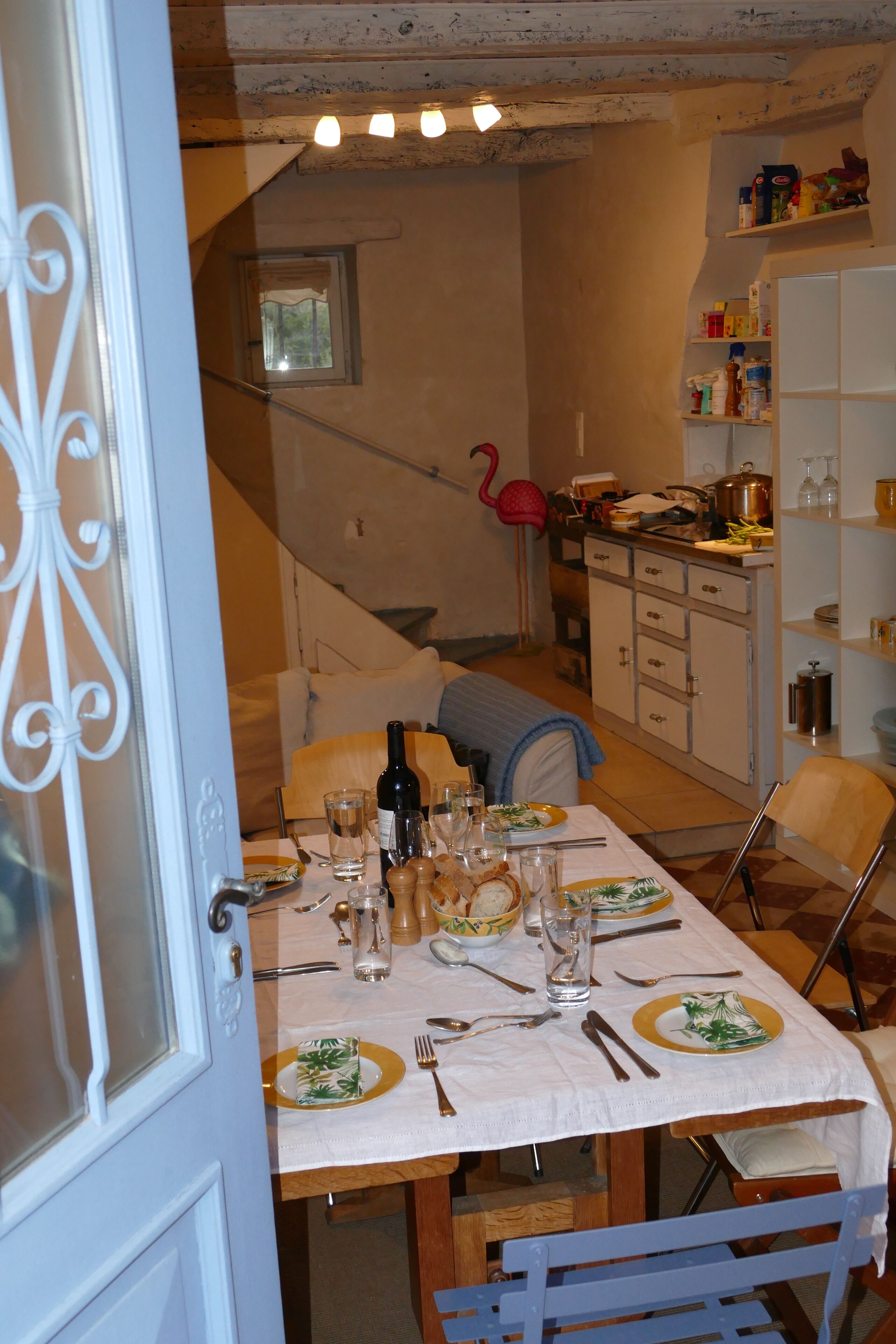 A dining area with a table set for four, a staircase, and a kitchen area with shelves and a flamingo decoration.