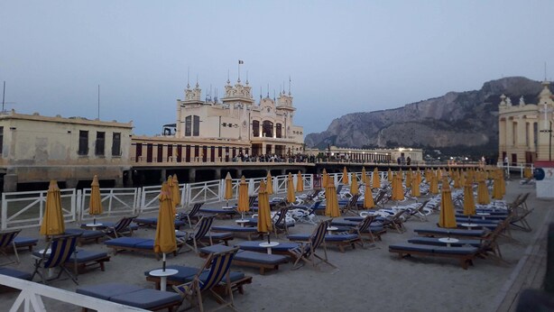 A beach with sun loungers and umbrellas, a large ornate building in the background, and a mountainous landscape.