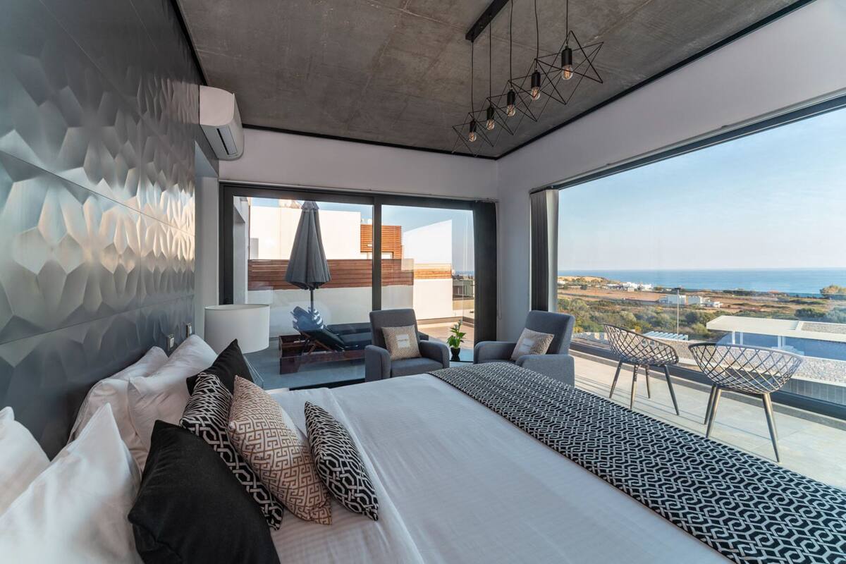 A modern bedroom with a large bed, a balcony with seating, and a view of the sea.