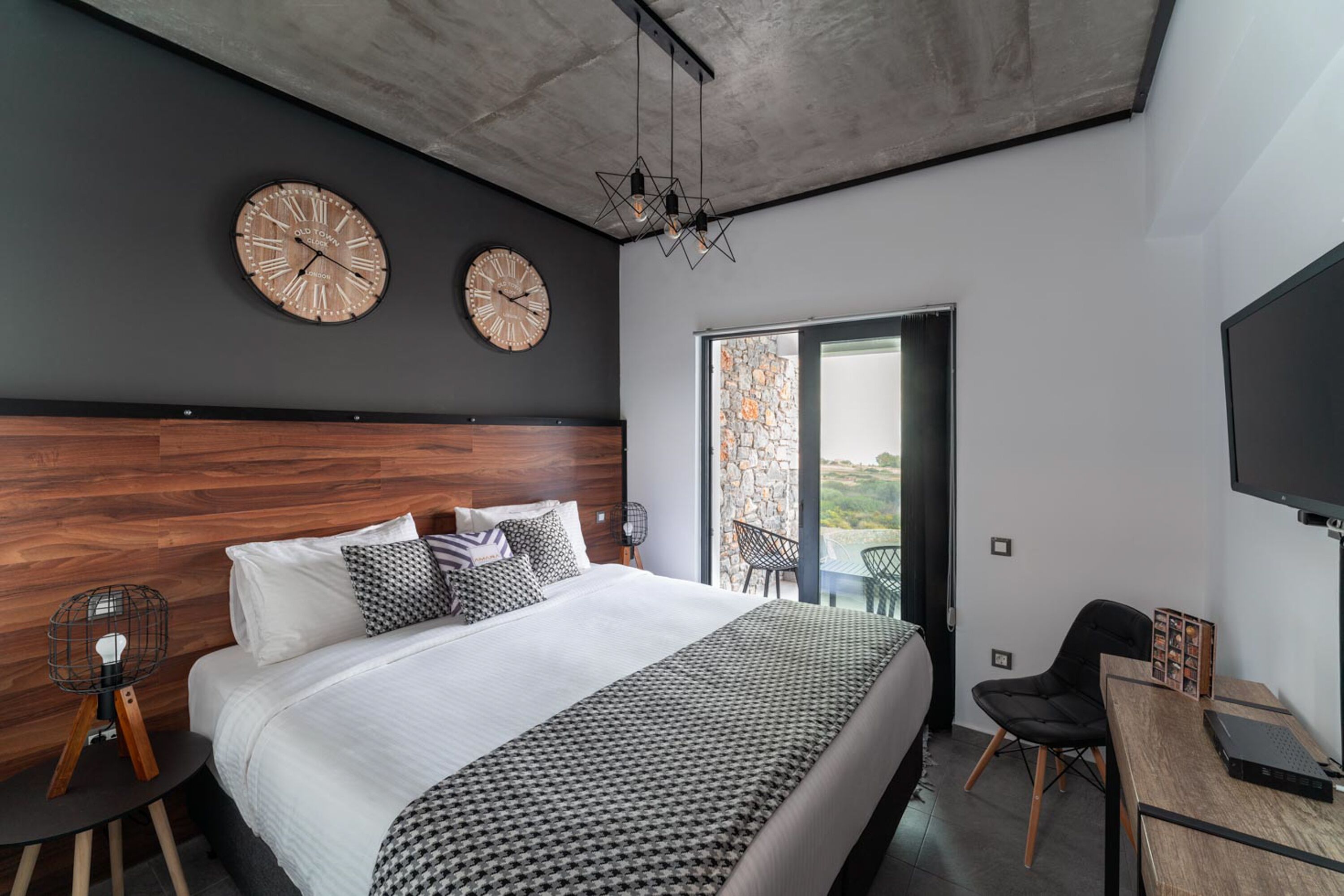 A modern bedroom with a large bed, two wall clocks, a ceiling light, and a view through a glass door.