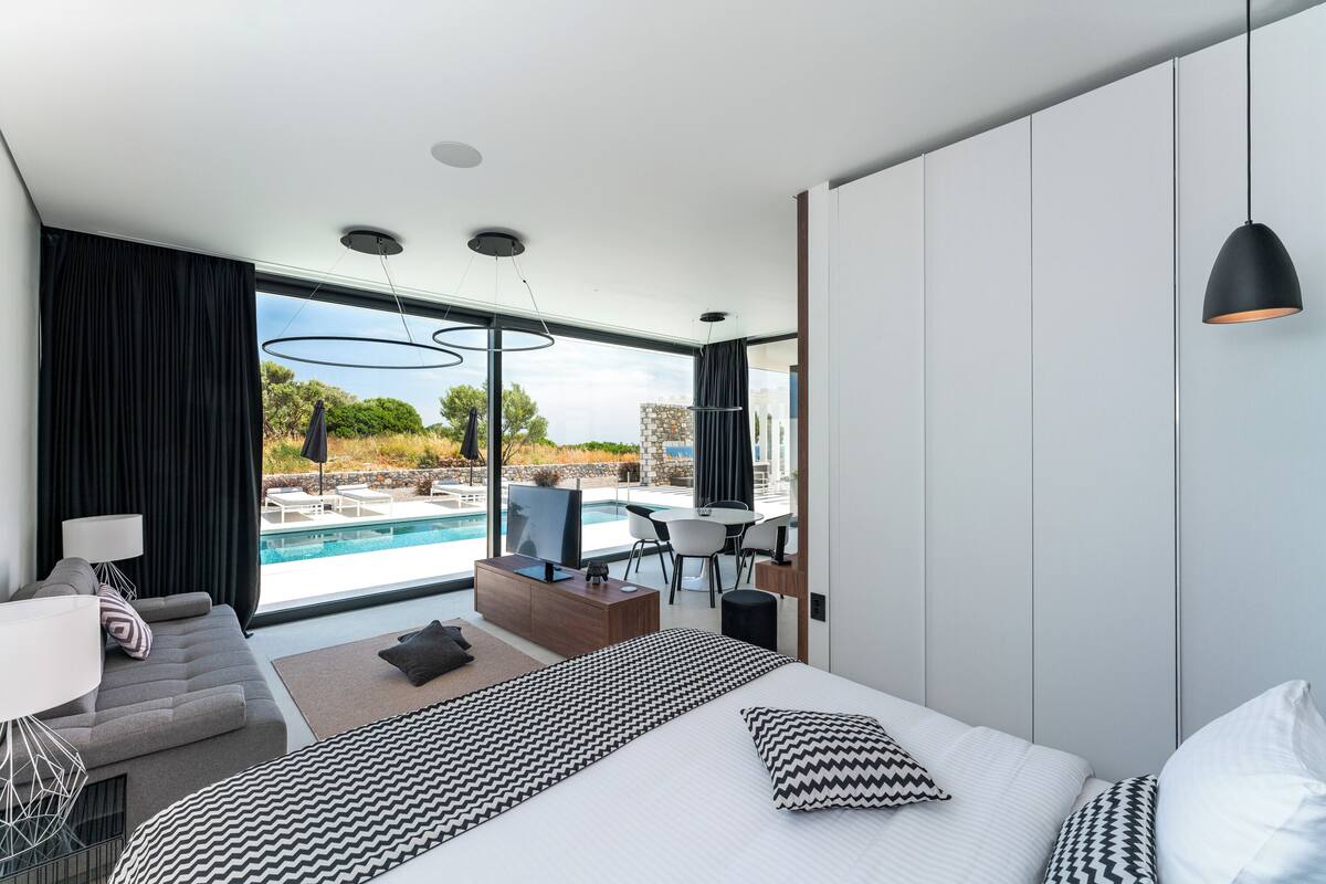 A modern hotel room with a large bed, a sofa, a desk, and a view of an outdoor swimming pool.