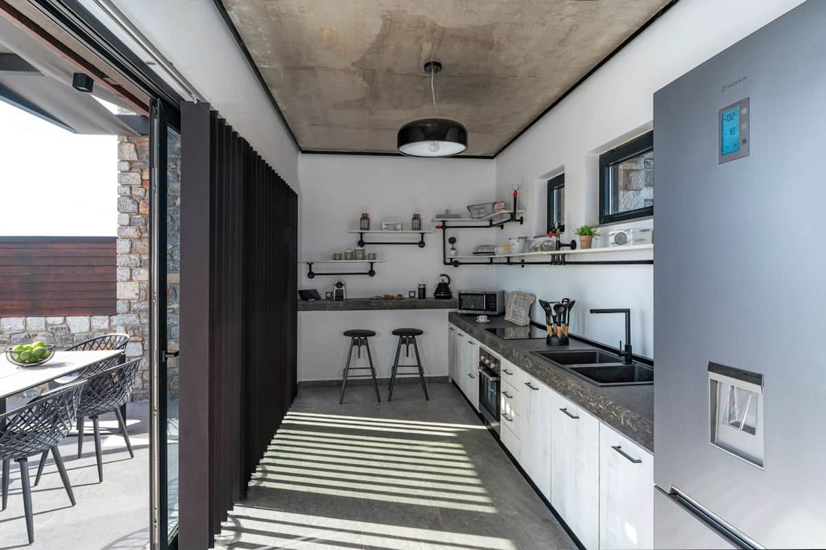 A modern kitchen with a built-in oven, a microwave, and a sink. There are two stools and a table with a bowl of fruit. The kitchen has a concrete ceiling and a window.