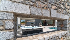 A modern outdoor hot tub area with a stone wall backdrop, a pool, and a contemporary building with large windows. - Amara Luxury villas (Rhodes)