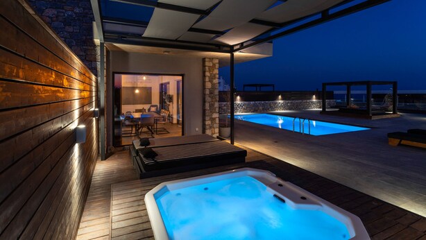 A modern outdoor pool area with a hot tub, wooden deck, and stone walls. - Amara Luxury villas (Rhodes)