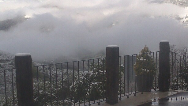 Een balkon met een metalen hekwerk, met uitzicht op een besneeuwd berglandschap, deels verborgen door wolken.