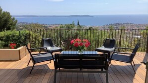A wooden deck with a table and four chairs, overlooking a coastal landscape with a view of the sea and distant hills.
