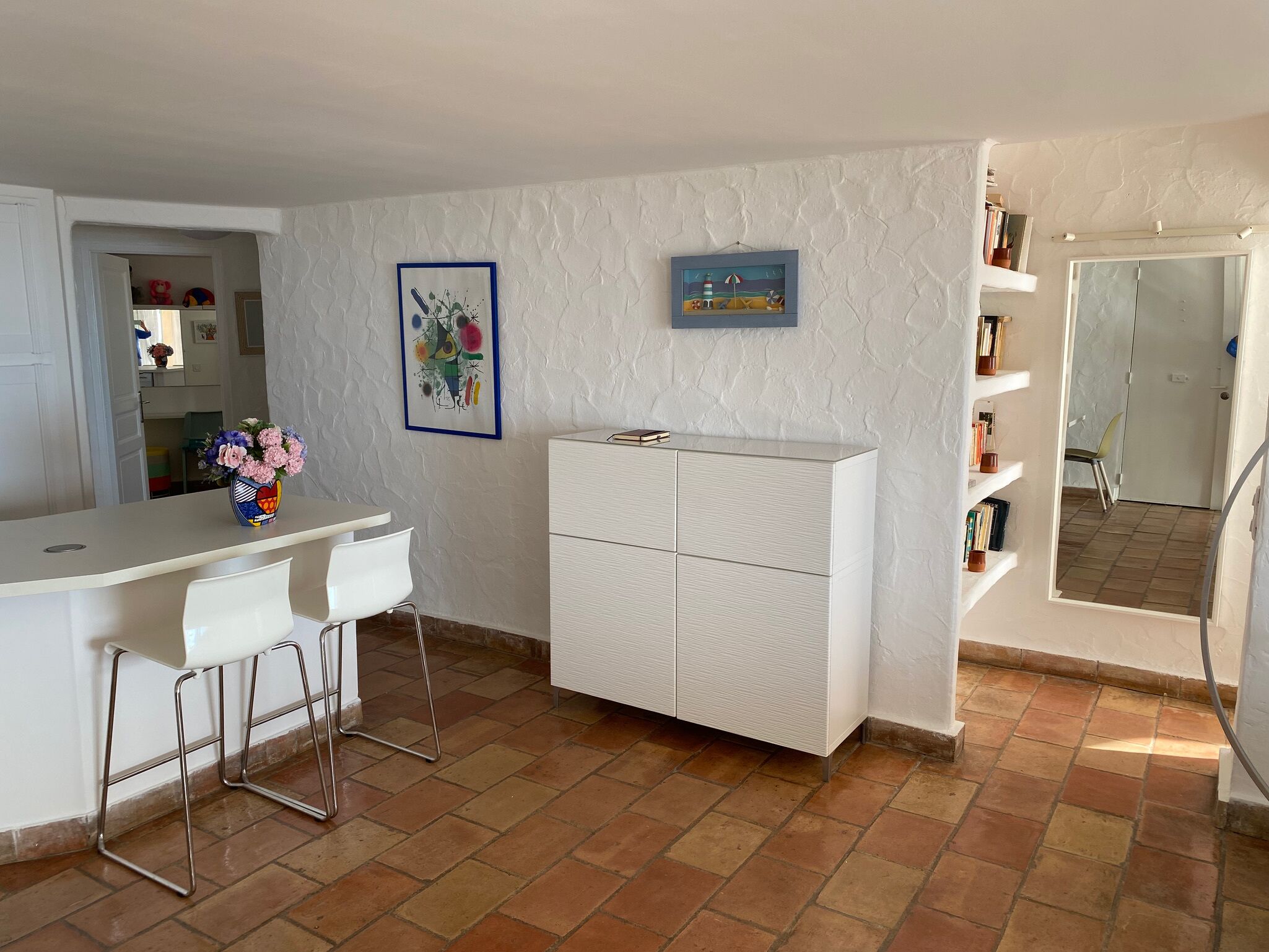 A compact kitchen with a white countertop, two bar stools, a white cabinet, and a small shelf with books.