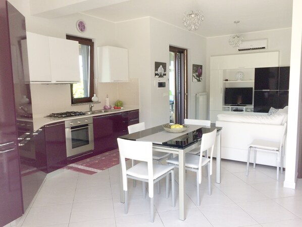 A modern kitchen with white cabinets, a dining table with chairs, and a living area with a sofa and TV.