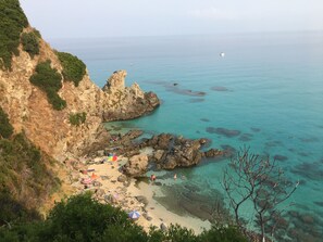 A coastal beach with rocky cliffs, clear turquoise water, and a sandy area with umbrellas.