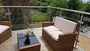 Un balcon extérieur doté de meubles en osier, d’une rambarde en verre et offrant une vue sur la verdure.