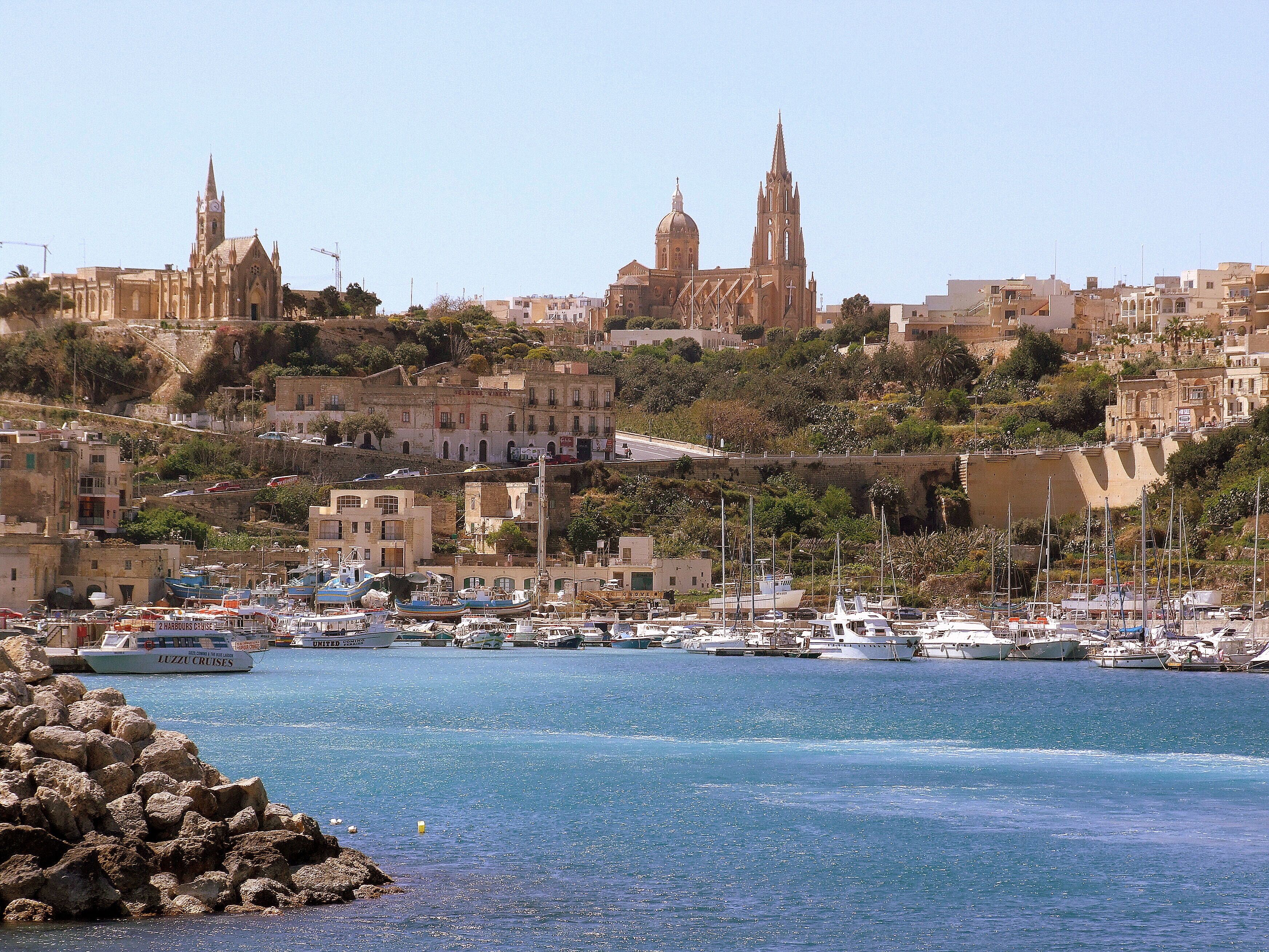A coastal cityscape with historic buildings, a marina with boats, and a prominent church.