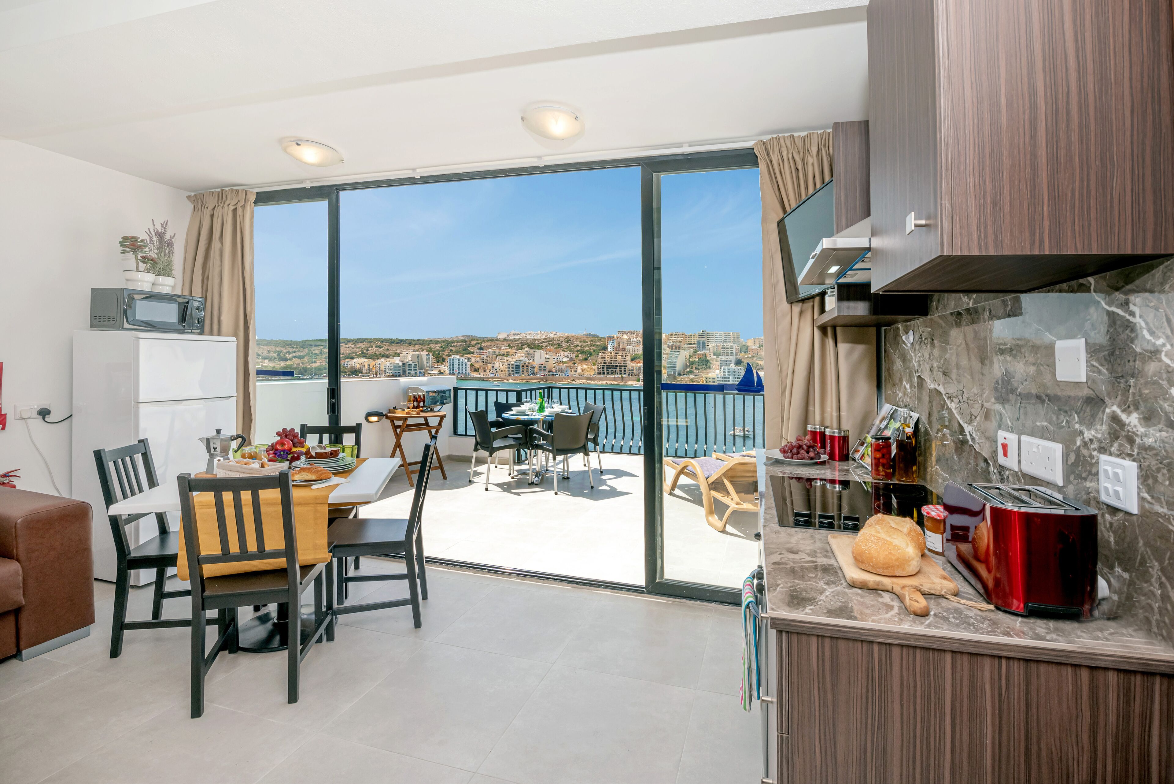 Cozinha moderna com uma ilha grande, área de jantar com mesa e cadeiras, e vista da cidade através de grandes portas de vidro deslizantes.