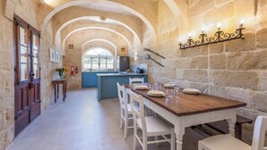 Comedor con paredes de piedra, una mesa de madera dispuesta para cuatro comensales, sillas blancas y una cocina al fondo.