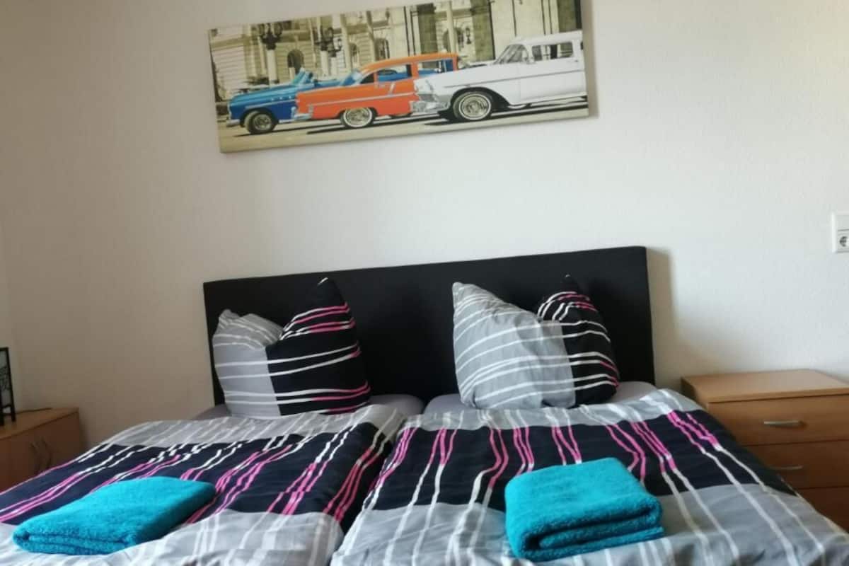 A neatly made bed with striped bedding and two towels, a nightstand with a lamp, and a wall-mounted picture of vintage cars.