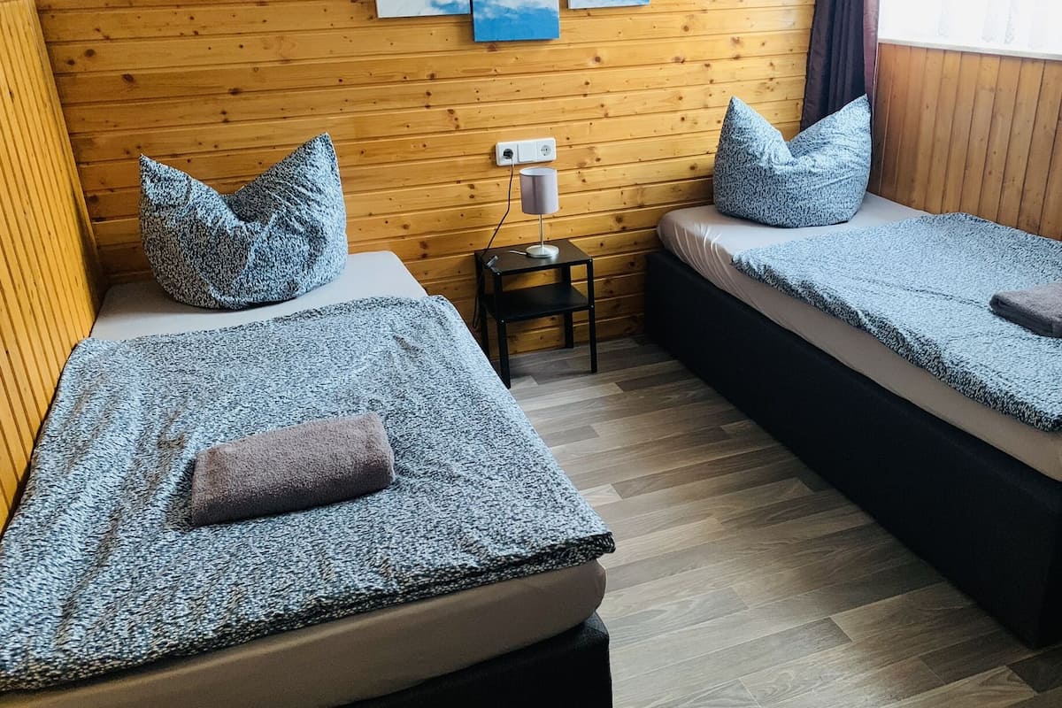 A room with two single beds, each with a pillow and a blanket. There is a small table with a lamp and a power outlet on the wall.