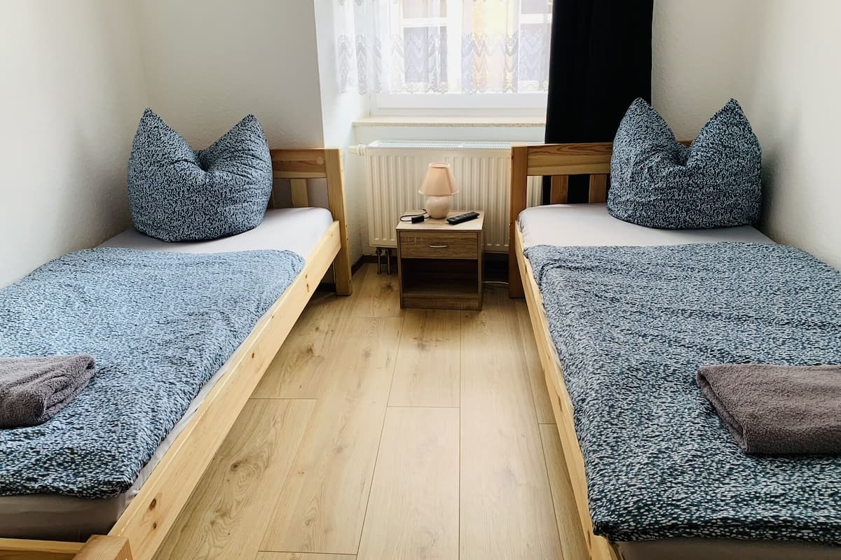 A small, cozy room with two single beds, each with a matching pillow and a blanket. There is a nightstand with a lamp and a remote control between the beds.