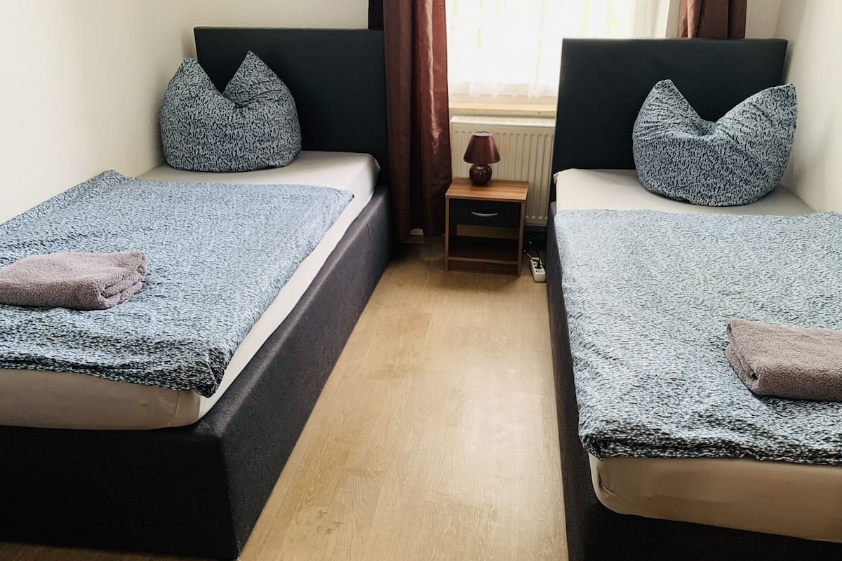 A room with two beds, each with a pillow and a blanket. There is a small table with a lamp between the beds.
