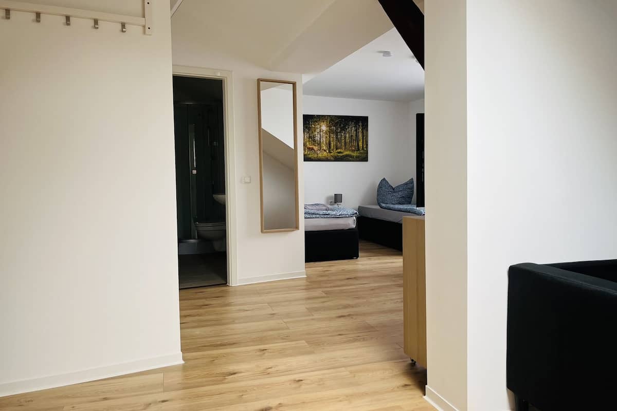 A modern interior with a wooden floor, a bed with a blue patterned bedspread, a painting of a forest, and a bathroom with a dark interior.