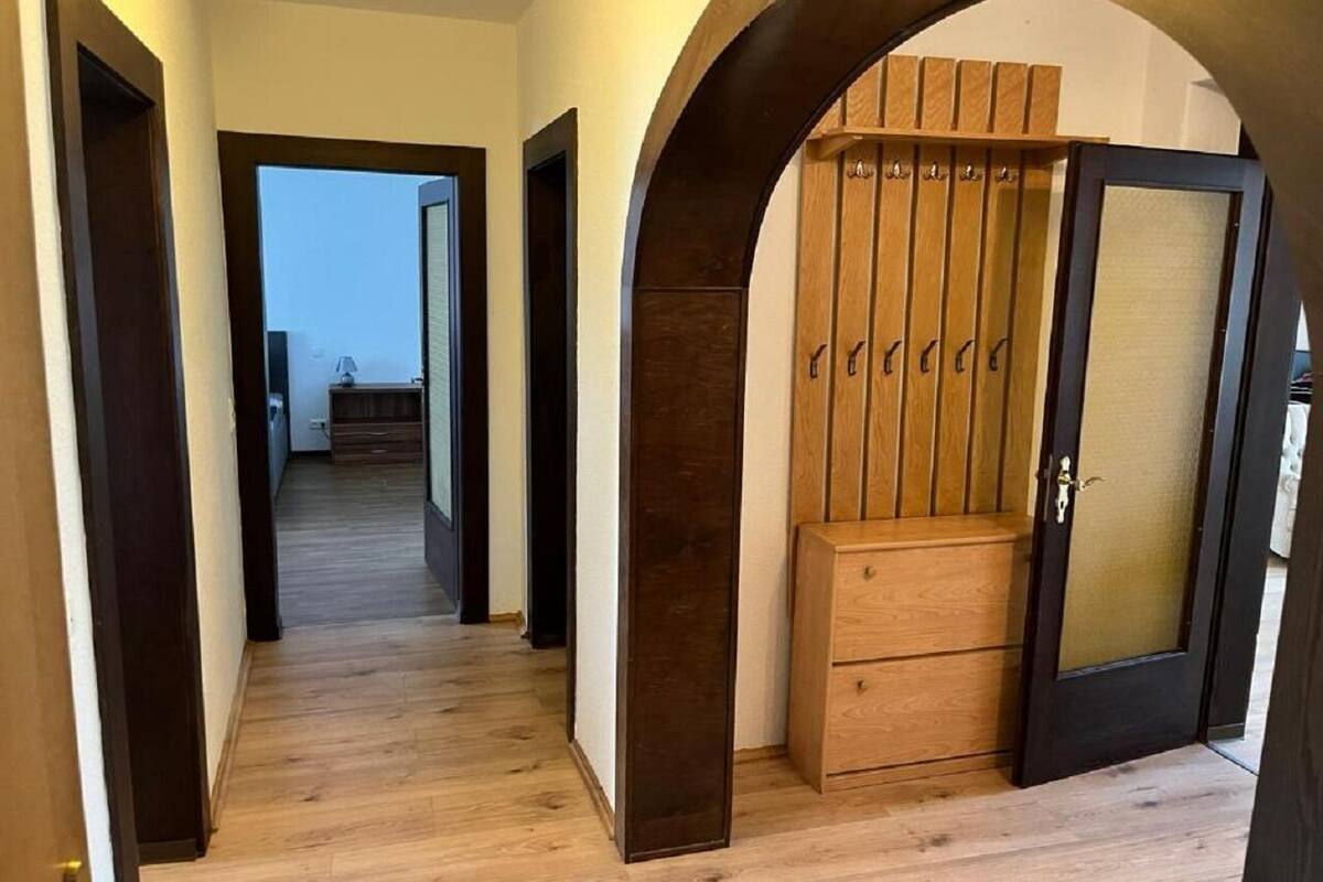 A hallway with wooden flooring, a dark wooden door with a glass panel, and a wooden cabinet with hooks.