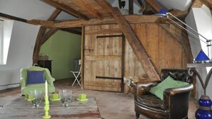 Quarto rústico com vigas de madeira aparentes, uma poltrona verde e uma poltrona de couro, além de uma porta de madeira.