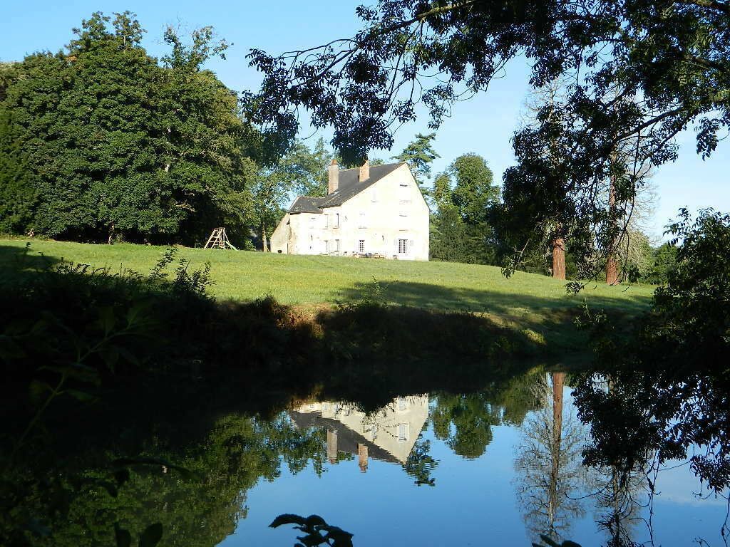 Une grande maison blanche qui se reflète dans un étang, entourée d’arbres et d’une zone herbeuse.