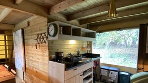 A wooden cabin interior with a kitchen area, wooden walls, a dining table, and a window with a view of trees.
