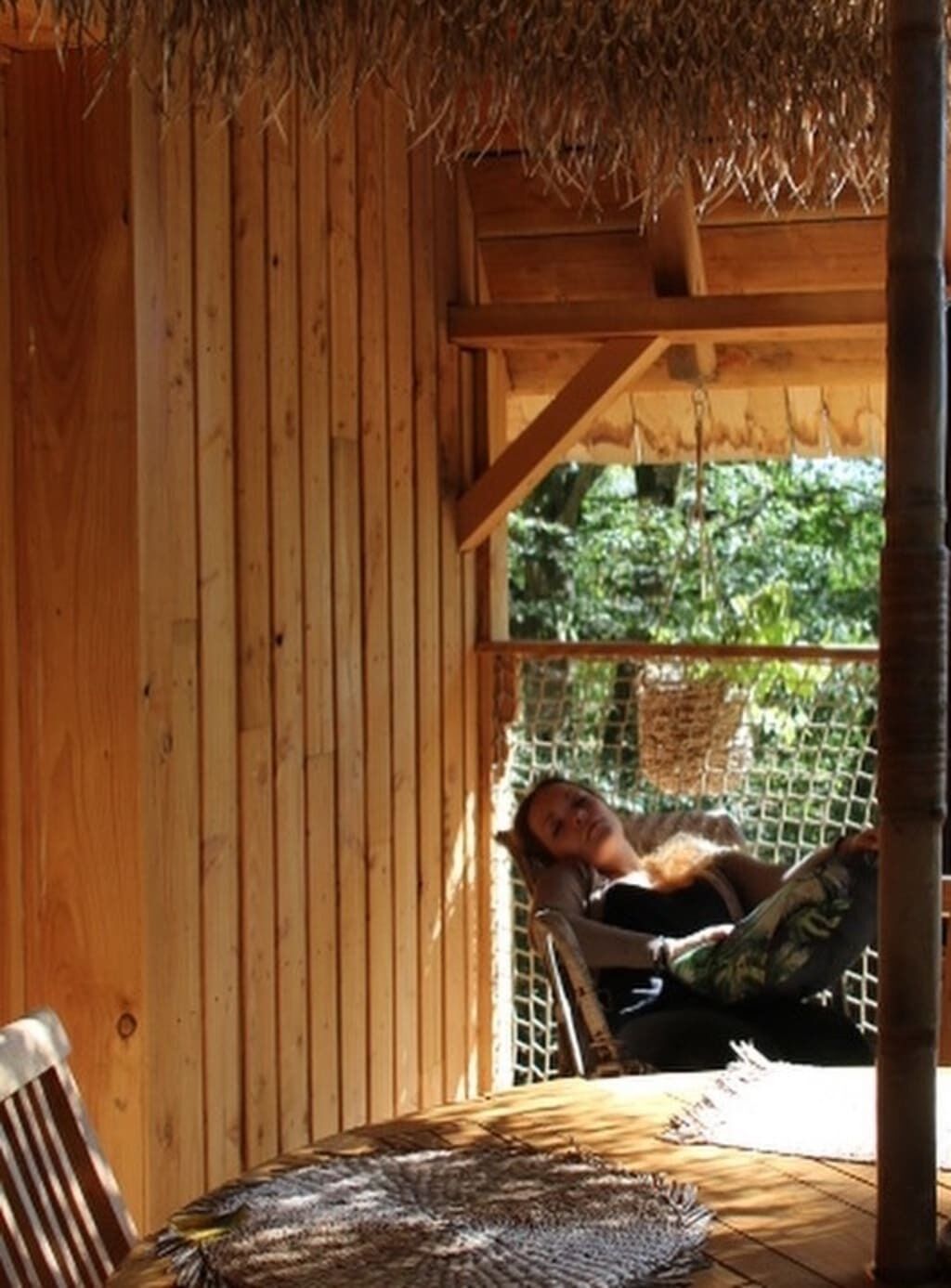 A wooden cabin with a thatched roof, a hammock, and a woven mat on the floor.
