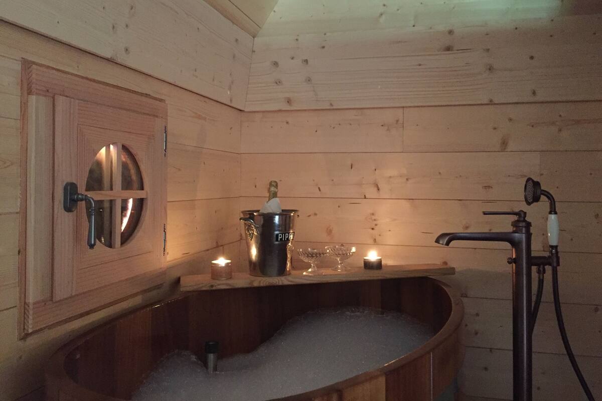 A wooden cabin interior with a wooden bathtub filled with water, a small candle, and a metal bucket on a wooden shelf.