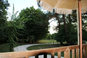 A wooden deck with a thatched umbrella, surrounded by trees and a gravel path. - Ecolodge Beauregard (Treigny-Perreuse-Sainte-Colombe)