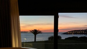 Un balcon offrant une vue sur un coucher de soleil sur la mer, un palmier et une ville lointaine.