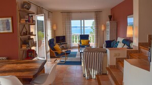A spacious living area with a large window offering a view of the sea, a wooden table, a sofa, and a television.