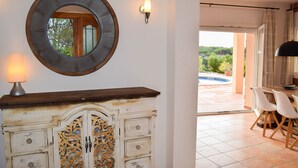 A room with a wooden cabinet, a wall-mounted mirror, a table with chairs, and a view of an outdoor area with a pool.