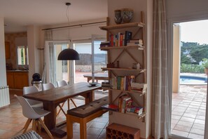 A dining area with a wooden table and chairs, a bookshelf with books, and a view of a swimming pool through an open door. - Villa with splendid sea view, 500 m. from the beach, Wifi, pool (Begur)