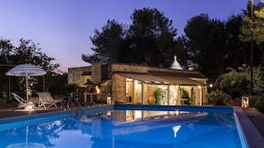 Une piscine avec une maison en pierre en arrière-plan, un fauteuil de relaxation et un parasol à rayures.