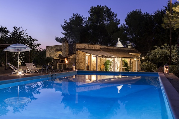 A pool with a stone house in the background, a lounge chair, and a striped umbrella.